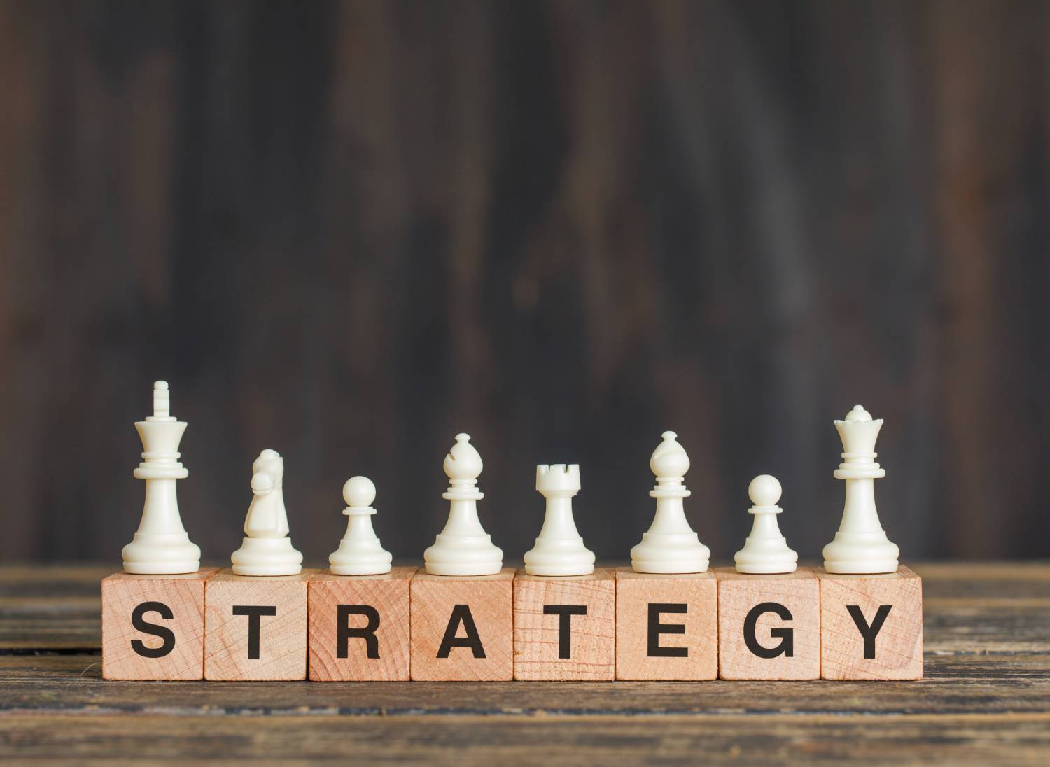 Chess pieces lined up behind blocks spelling "STRATEGY" on a wooden surface.